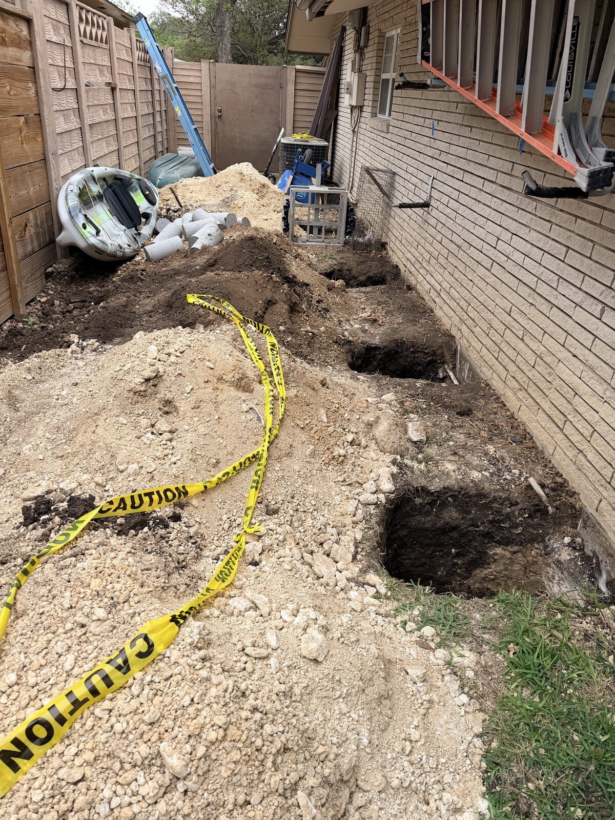 Side-yard foundation repair in progress with multiple pier pits and caution tape protecting the area