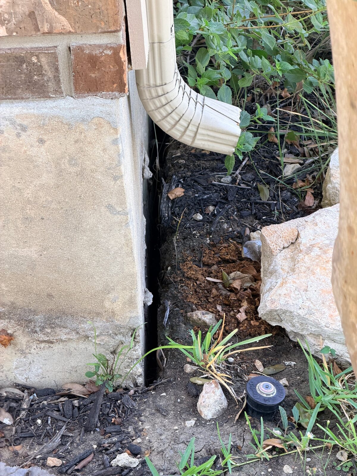 Dark open gap between dry soil and a concrete grade beam beside a downspout and a sprinkler head