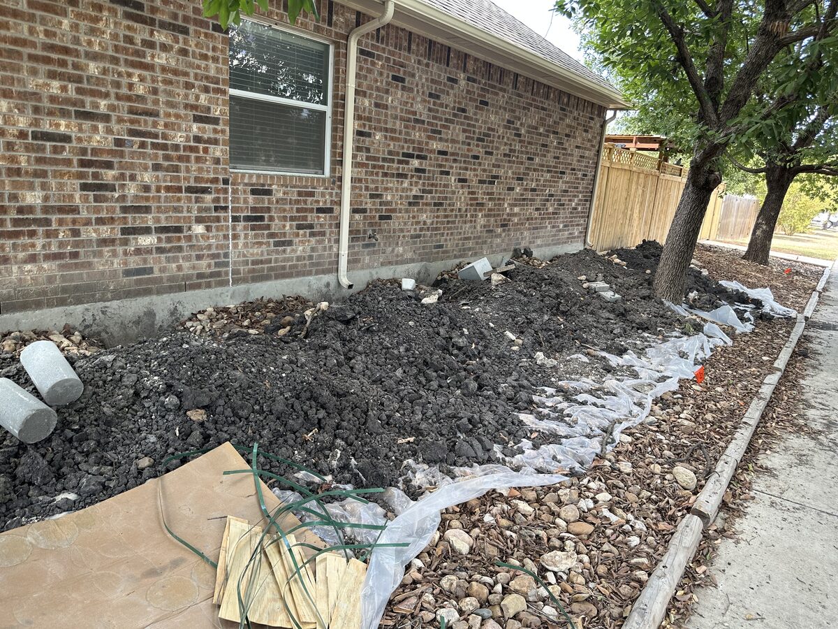 Excavated soil staged on landscape fabric along a brick home, awaiting backfill after pier installation