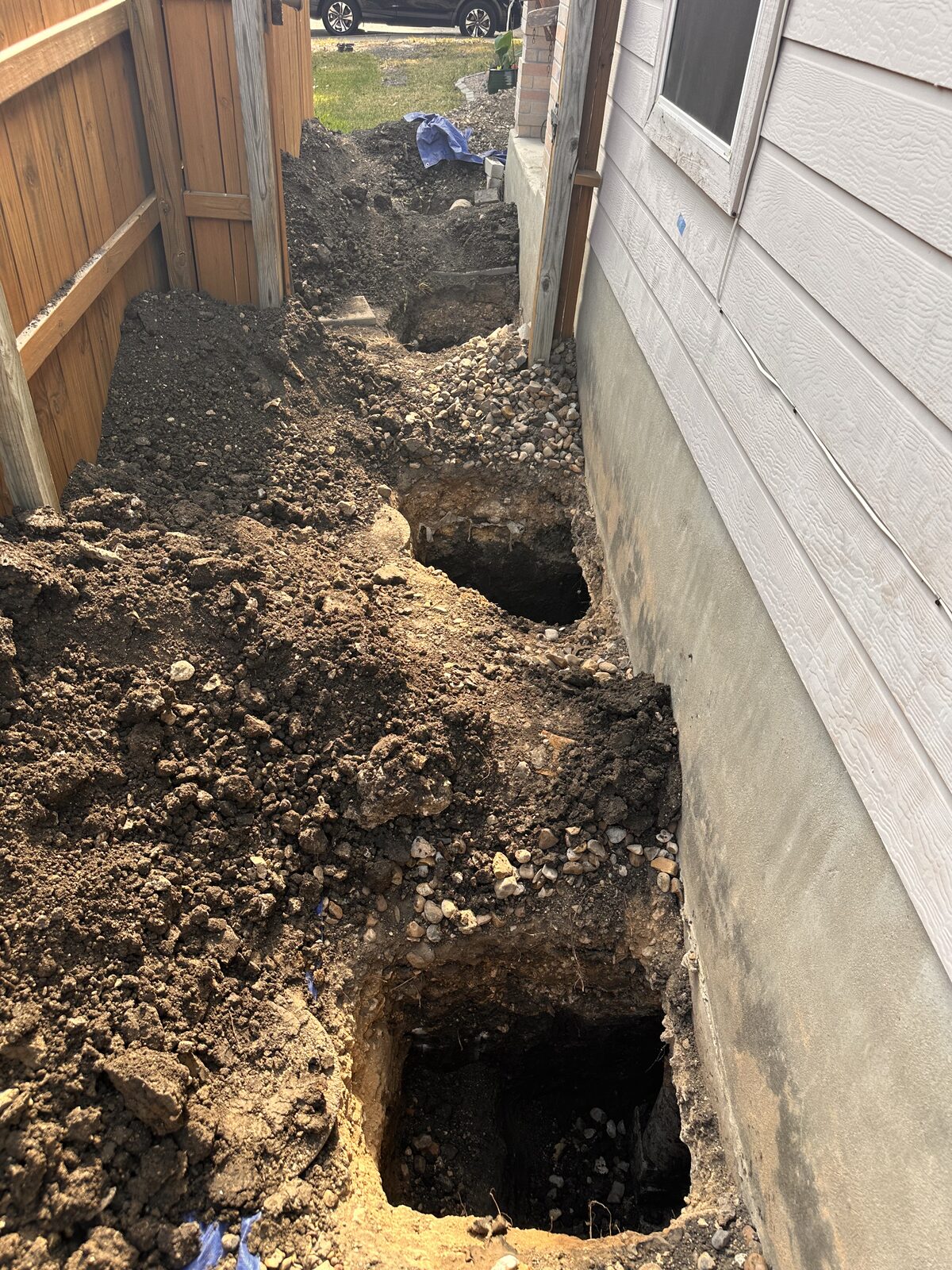 Square pier holes excavated along a concrete stem wall in a narrow fenced side yard