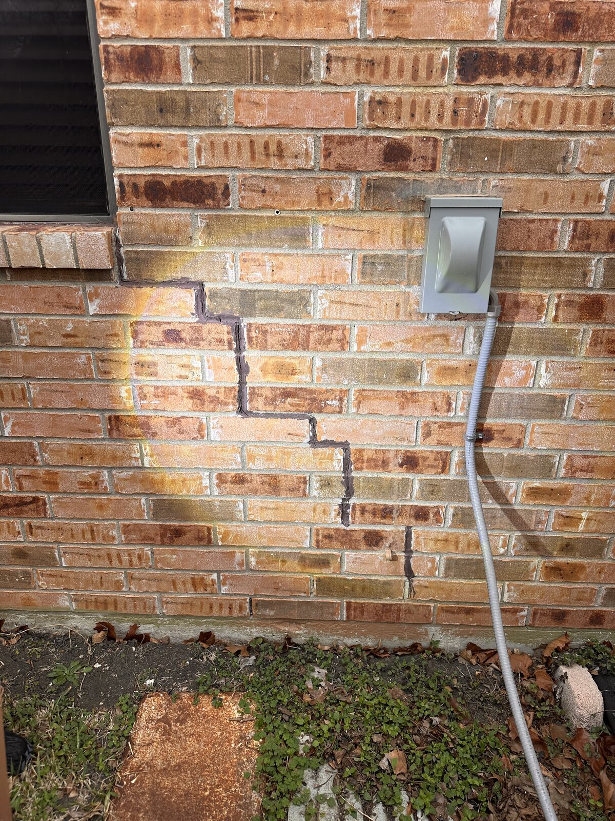 Stair-step crack in brick veneer below a window beside electrical conduit, a classic indicator of foundation settlement