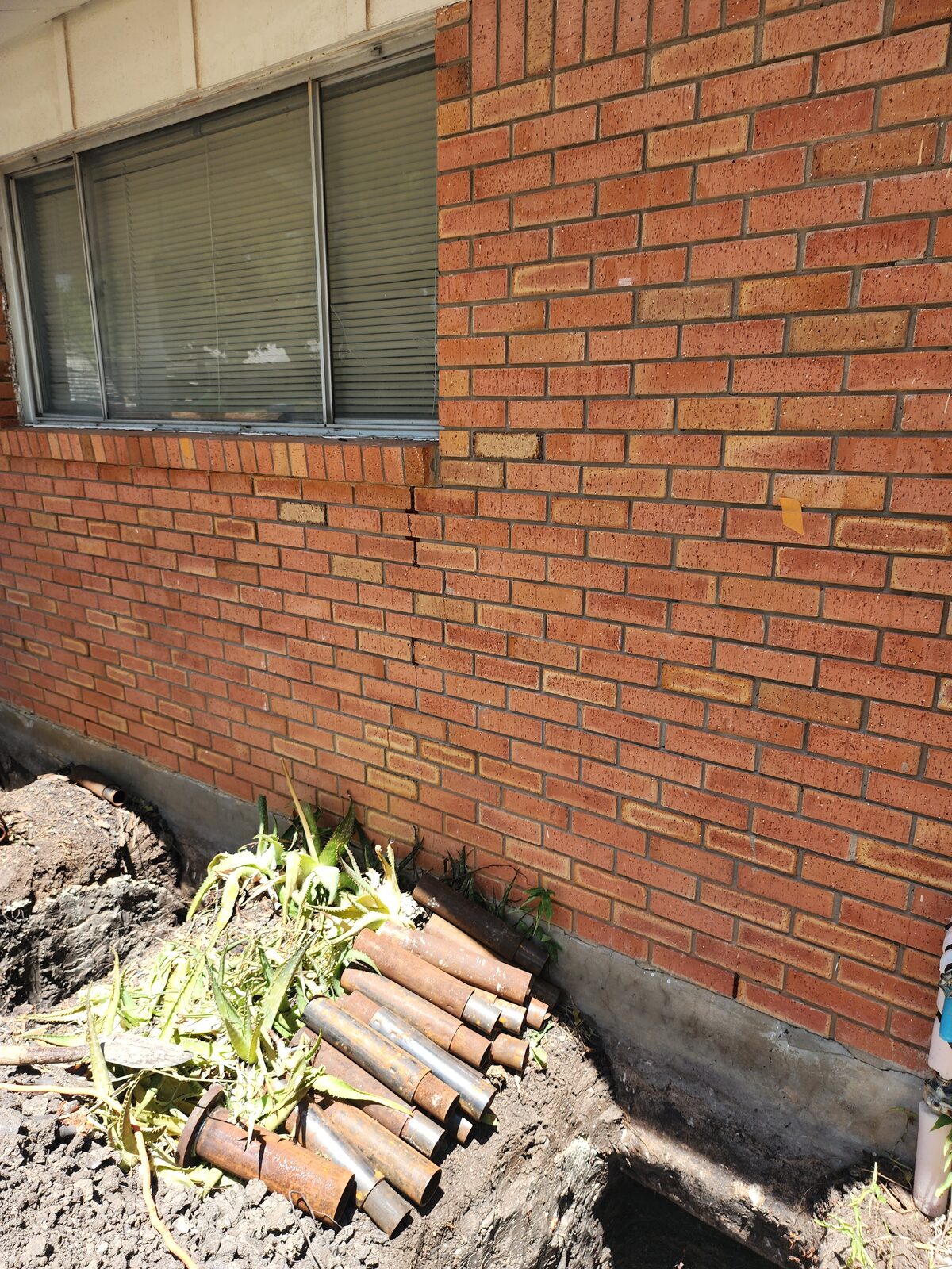 Steel pier pipe sections staged in an excavation beside a brick wall, ready for hydraulic pier driving
