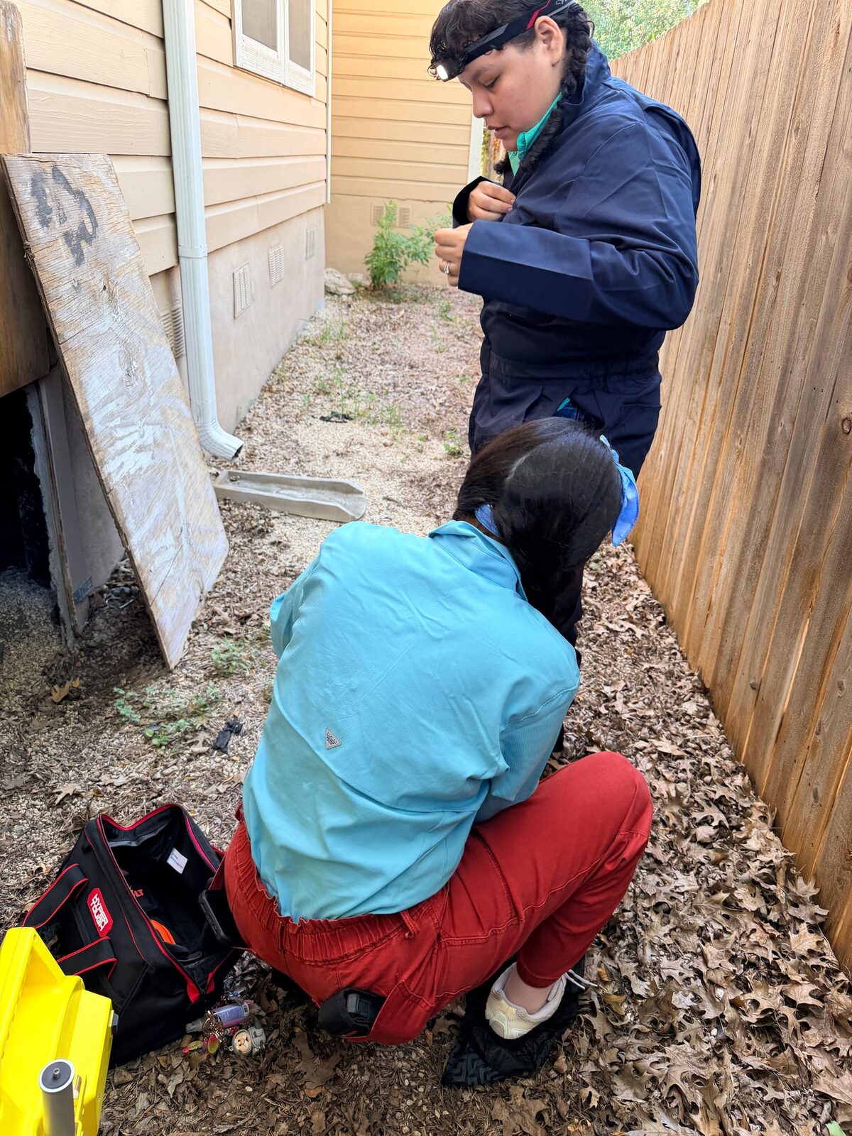 Crawl space access panel leaned against a house while two crew members get ready to go under
