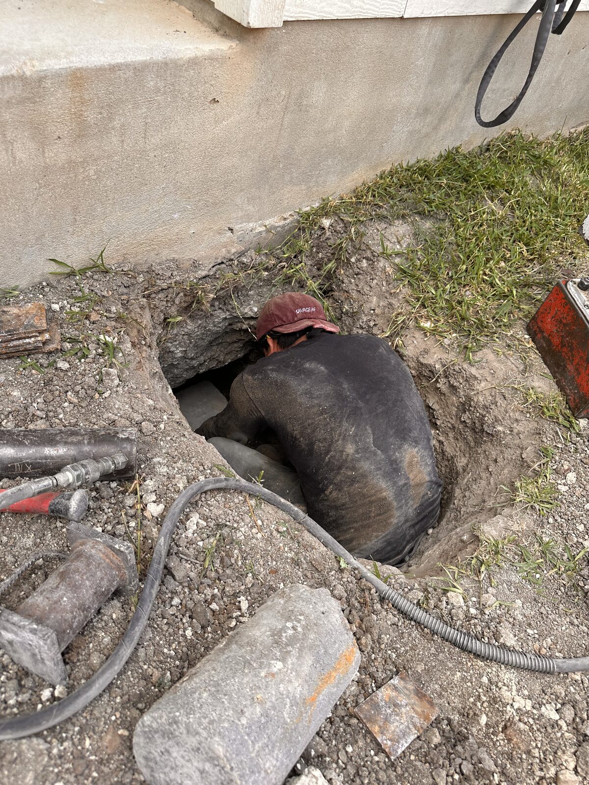 Foundation technician leaning into a hand-dug pit beside a slab, with hydraulic hoses and piling staged nearby