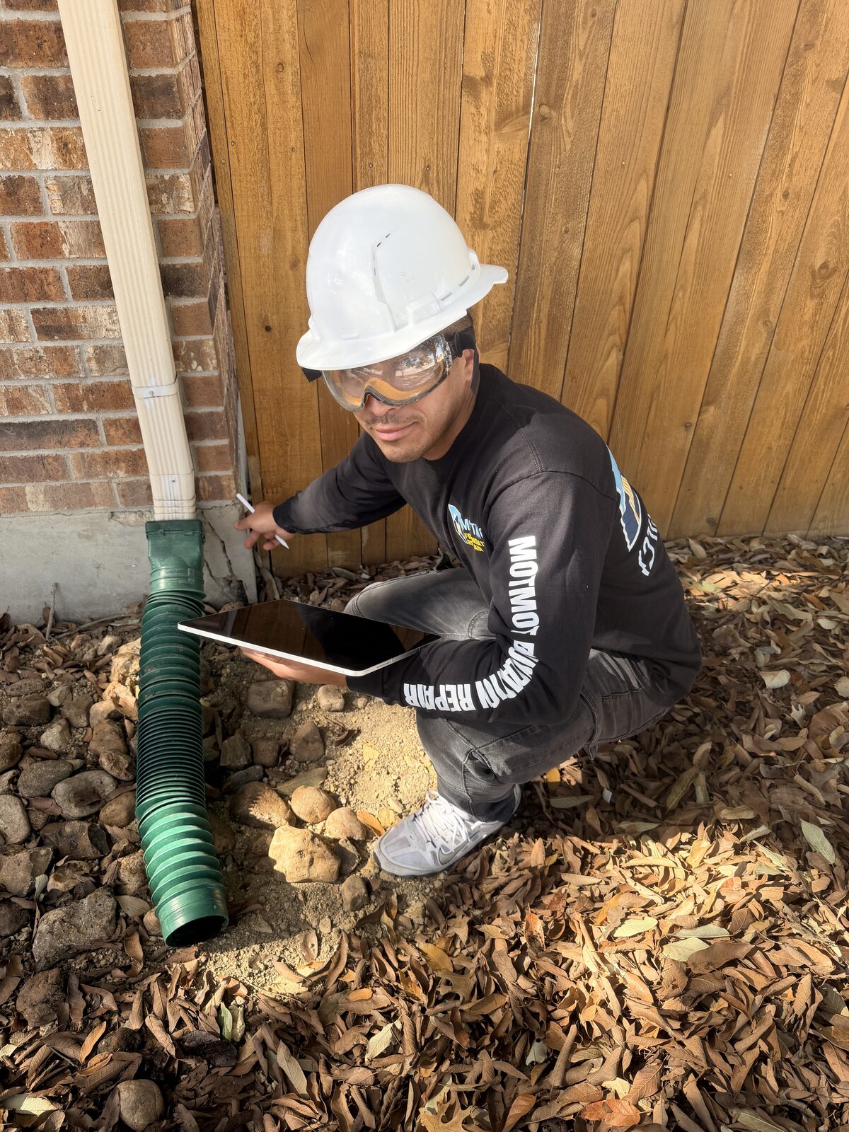 Technician inspecting a buried downspout drain extension beside a foundation