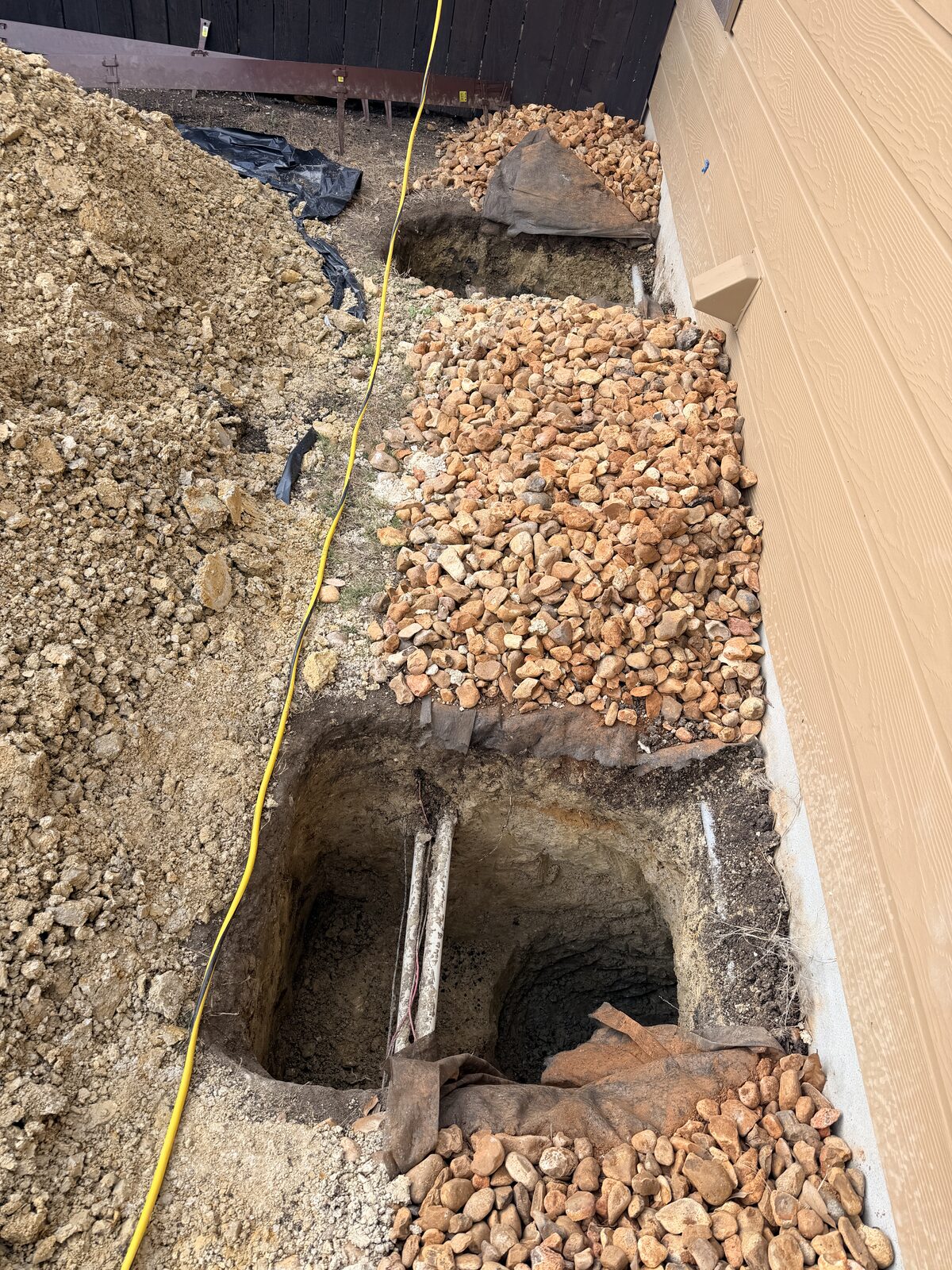 Two pier holes excavated along an exterior foundation wall, laid out with a tape measure for pier placement