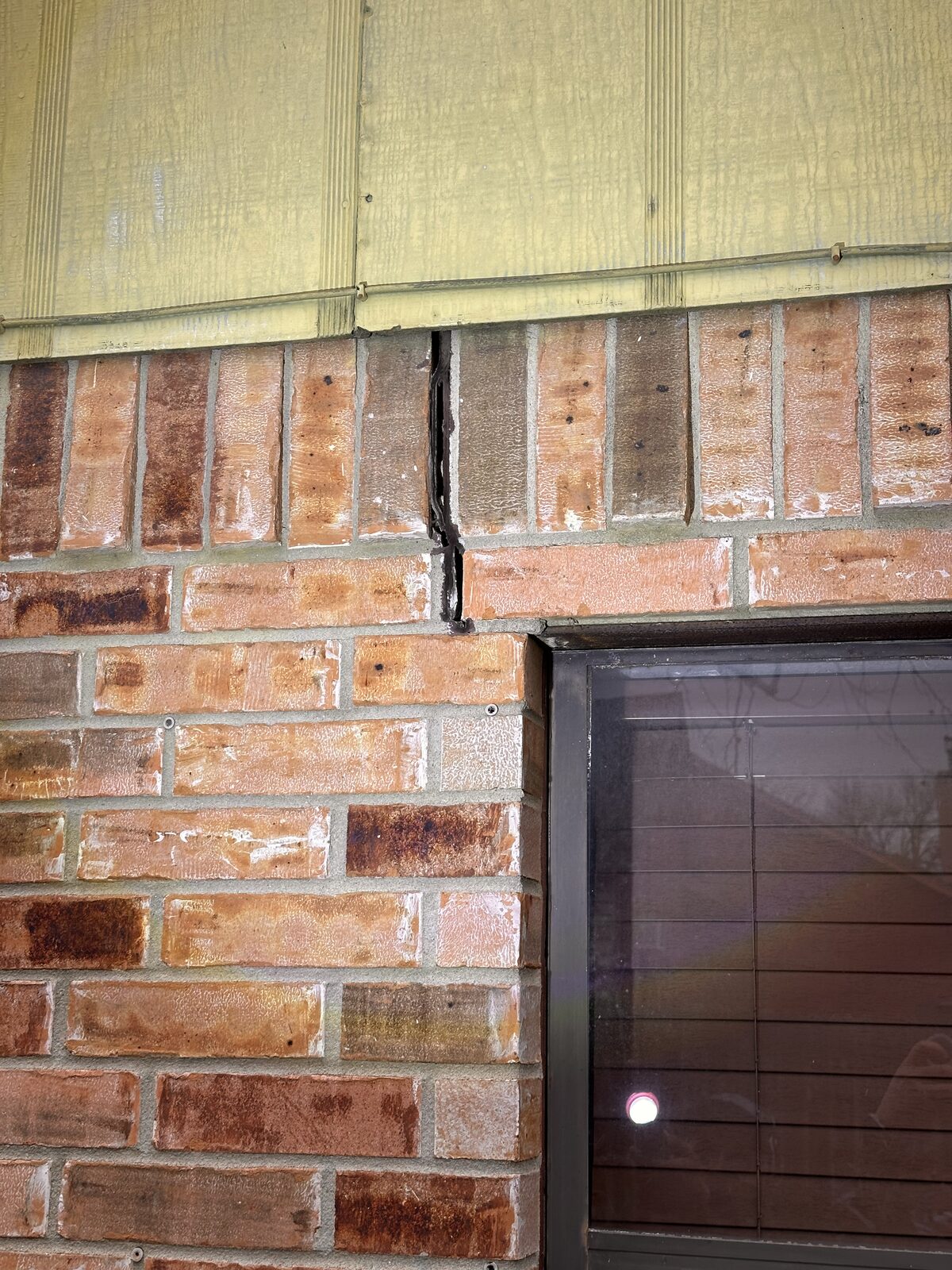 Vertical crack running from siding down through brick veneer beside a window, a sign of foundation settlement