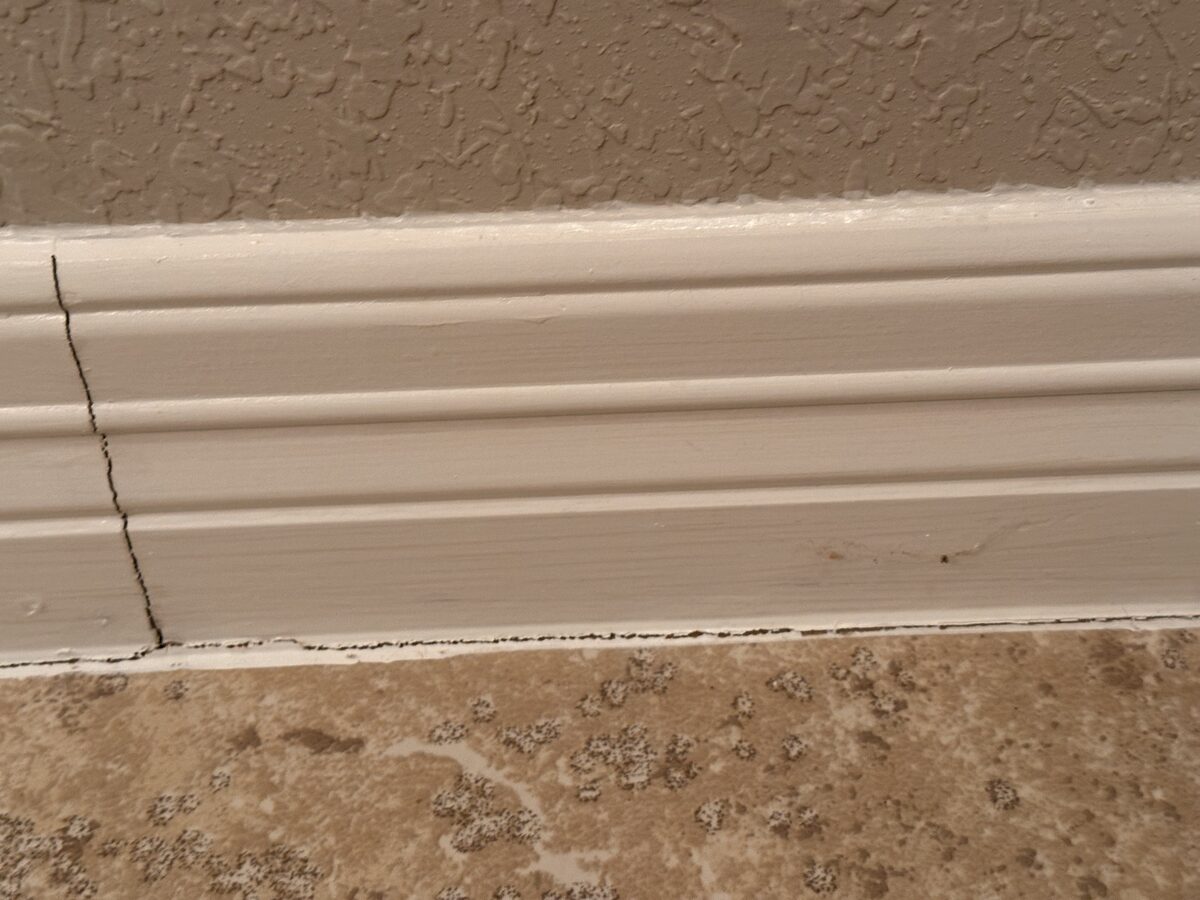 Vertical crack running through baseboard trim with a gap at the tile floor below, from slab movement