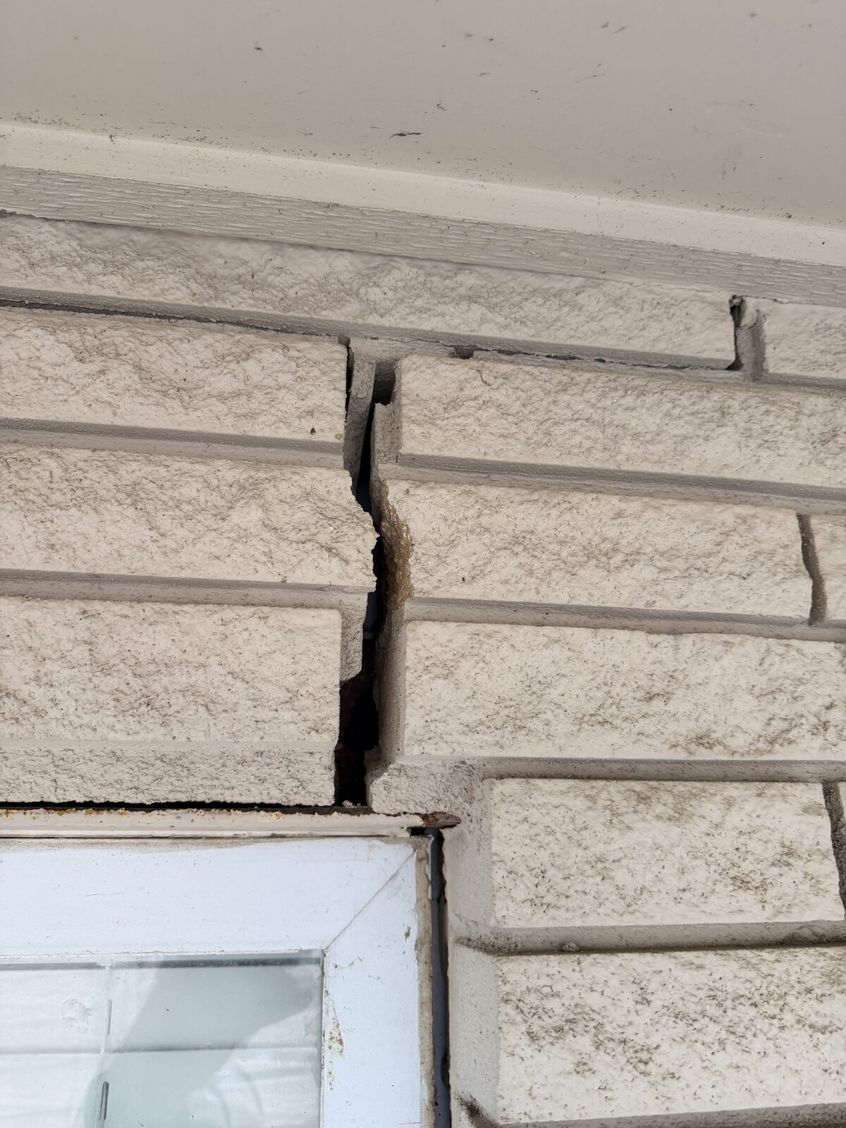 Wide open crack through pale stone veneer running down from the soffit to the top corner of a window